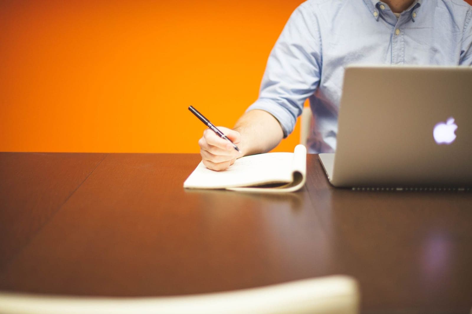Adult writing notes in a notebook beside a laptop on a wooden desk