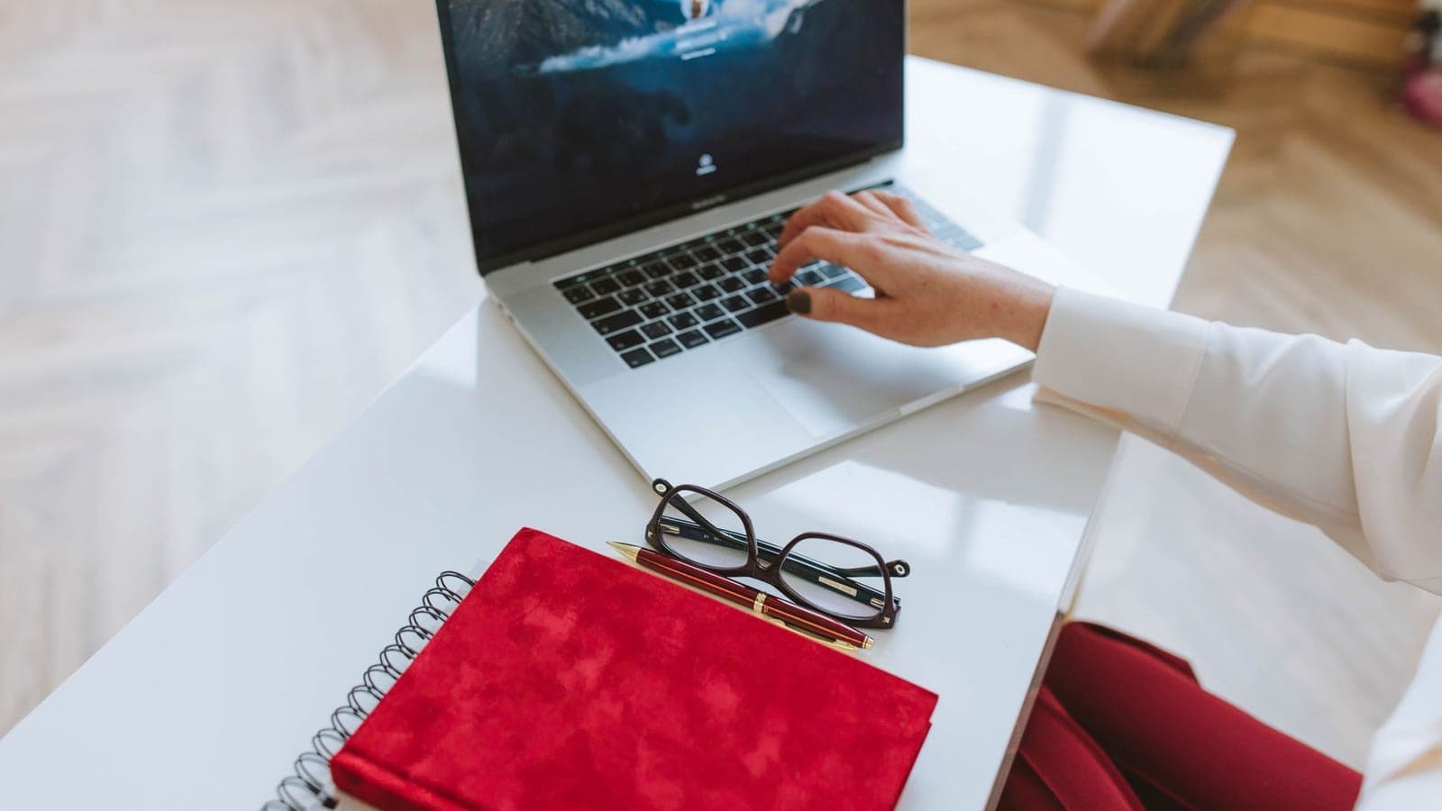Person working on a laptop at a white desk with a notebook and glasses